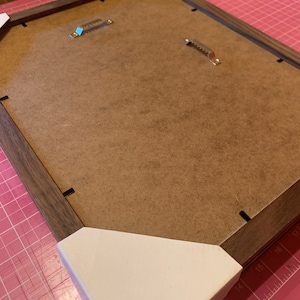 May include: The back of a brown wooden picture frame with white corner protectors. The frame has two metal hanging hooks.