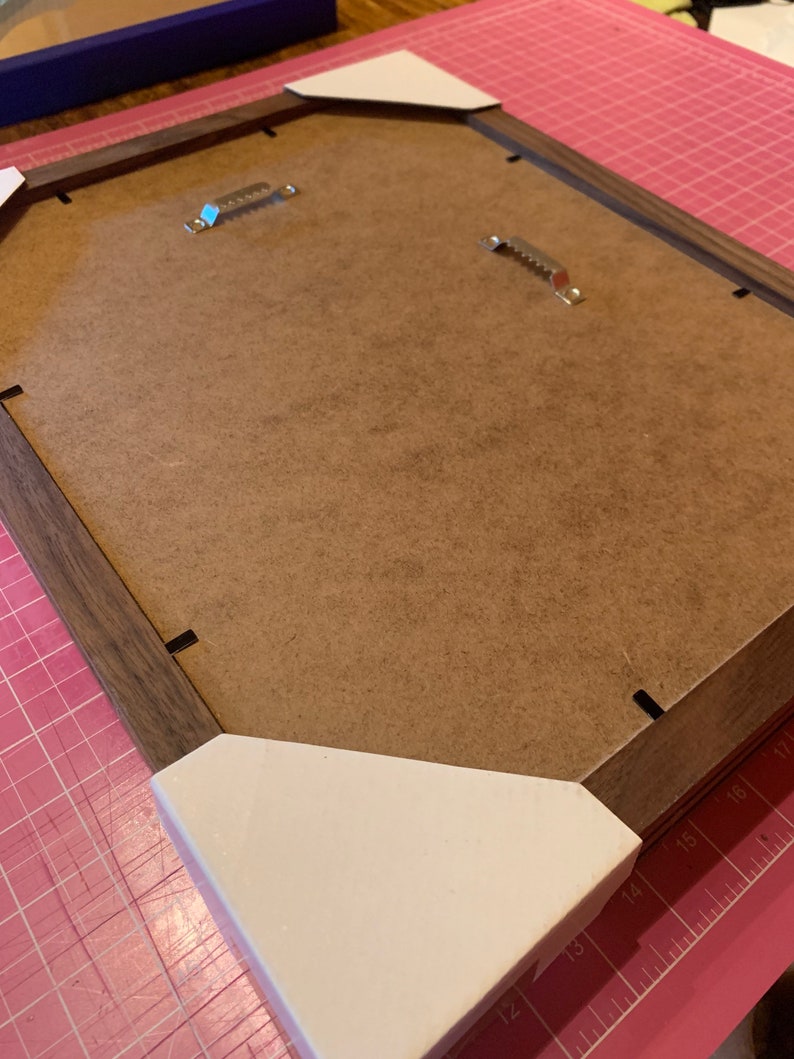 May include: The back of a brown wooden picture frame with white corner protectors. The frame has two metal hanging hooks.