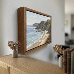 May include: A landscape painting of a coastal scene, framed in natural wood. The artwork depicts a beach, cliffs, and the sea. The painting is mounted on a white wall. The color palette includes blues, greens, and beige, creating a serene atmosphere.