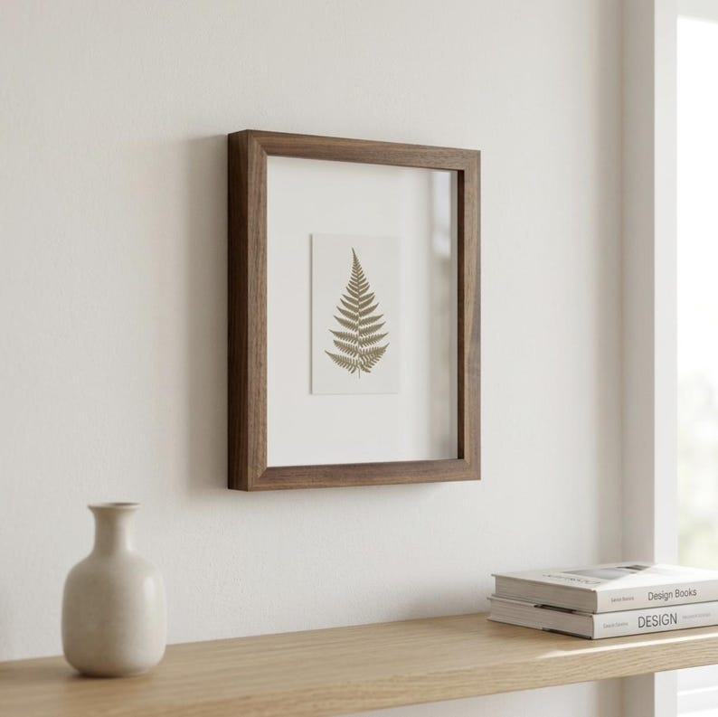 May include: A framed fern print with a dark brown wooden frame. The fern is a light gold color and is centered on a white background. The frame is rectangular and mounted on a white wall. A small vase and books are on a wooden shelf below.