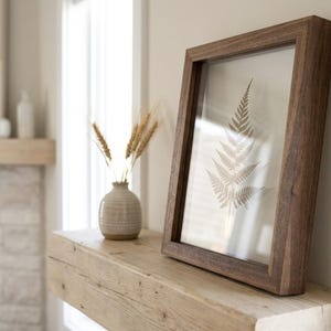 May include: A framed botanical print featuring a fern leaf, displayed on a wooden mantel. A small vase with wheat stalks sits nearby. The frame is dark brown, and the overall aesthetic is neutral and natural.