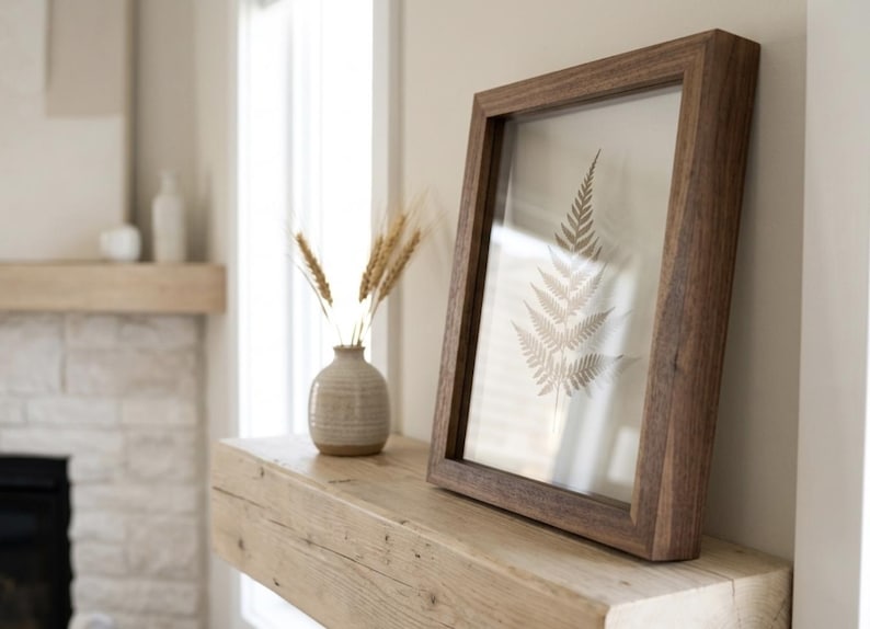 May include: A framed botanical print featuring a fern leaf, displayed on a wooden mantel. A small vase with wheat stalks sits nearby. The frame is dark brown, and the overall aesthetic is neutral and natural.