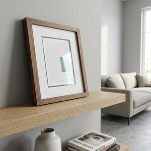 May include: A framed picture with a brown wooden frame, white mount, and teal detail. The artwork is displayed on a light-coloured wooden shelf against a grey wall. A sofa is visible in the background.