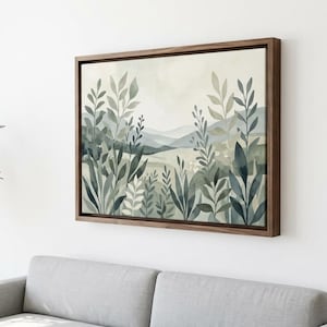 May include: A framed watercolour painting depicting a landscape with stylised plants and mountains in shades of green, blue, and beige. The artwork is displayed on a white wall above a grey sofa. The frame is made of dark wood.