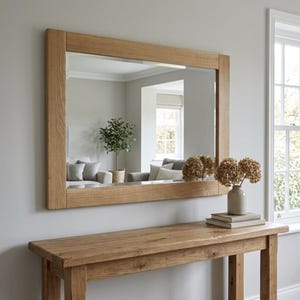 May include: A rectangular mirror with a light oak frame is mounted above a matching wooden console table. The mirror reflects a room with a sofa, a plant, and a window. A vase of dried flowers and books sit on the table.