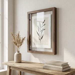 May include: A framed botanical print featuring a minimalist illustration of a leafy branch in shades of gray and black. The artwork is encased in a dark wood frame with a clear glass cover, displayed on a light-colored wall. A vase with dried plants and a stack of books are on a wooden table.