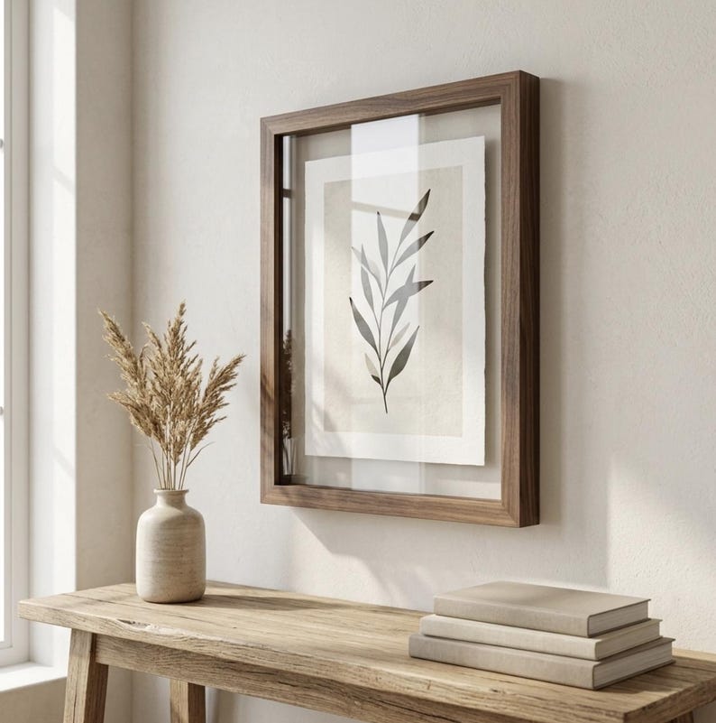 May include: A framed botanical print featuring a minimalist illustration of a leafy branch in shades of gray and black. The artwork is encased in a dark wood frame with a clear glass cover, displayed on a light-colored wall. A vase with dried plants and a stack of books are on a wooden table.