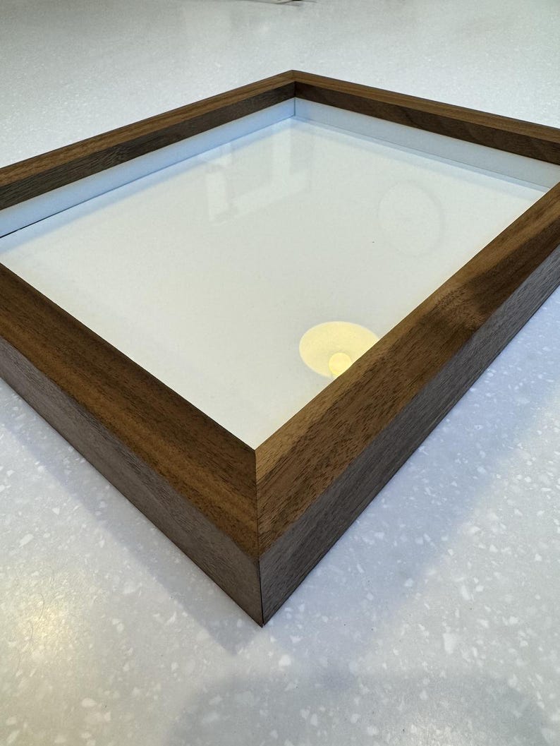 May include: A wooden picture frame with a clear glass front and a white interior. The frame is made of dark brown wood and has a simple, modern design. The frame is empty and ready to display a photo or artwork.