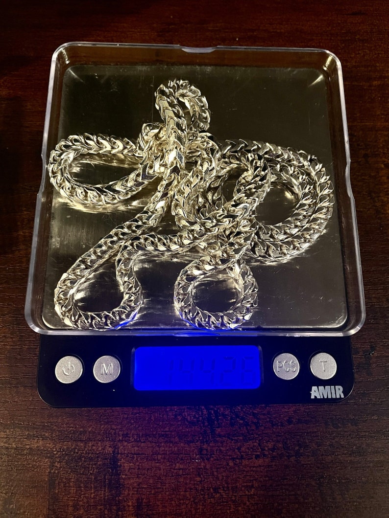 May include: A gold chain necklace on a digital scale, the scale reads 104.21. The scale is black with a blue digital display and has buttons labeled "M", "PCS", and "T". The scale is labeled "AMIR".