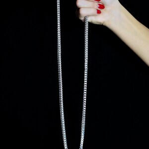 May include: A silver chain necklace with a box chain design. The chain is held in a hand with red nail polish.