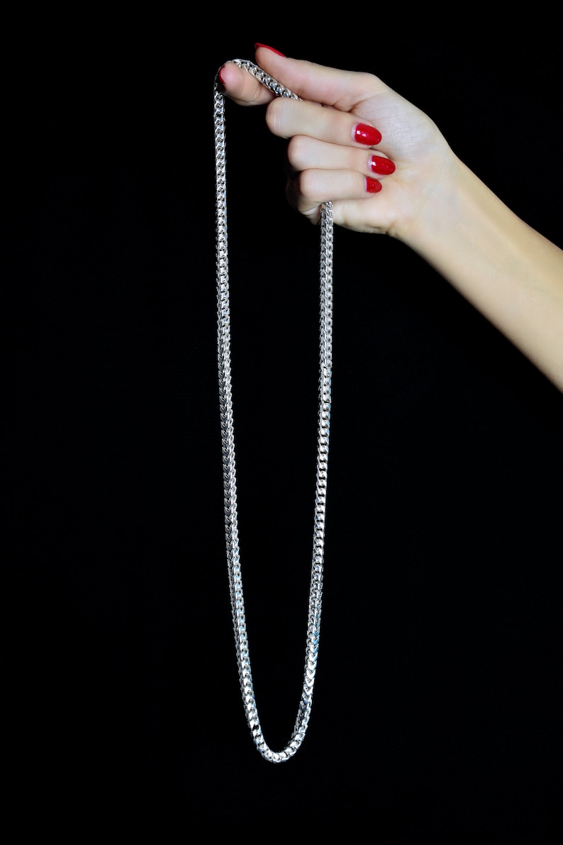 May include: A silver chain necklace with a box chain design. The chain is held in a hand with red nail polish.