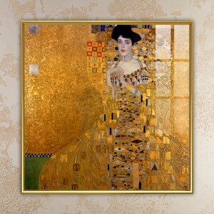 Op de afbeelding: Een ingelijste print van een schilderij van Gustav Klimt. Het kunstwerk toont een vrouw in een gouden jurk en hoed, tegen een gouden achtergrond. Het schilderij heeft ingewikkelde patronen en details, met een focus op goud- en aardetinten.