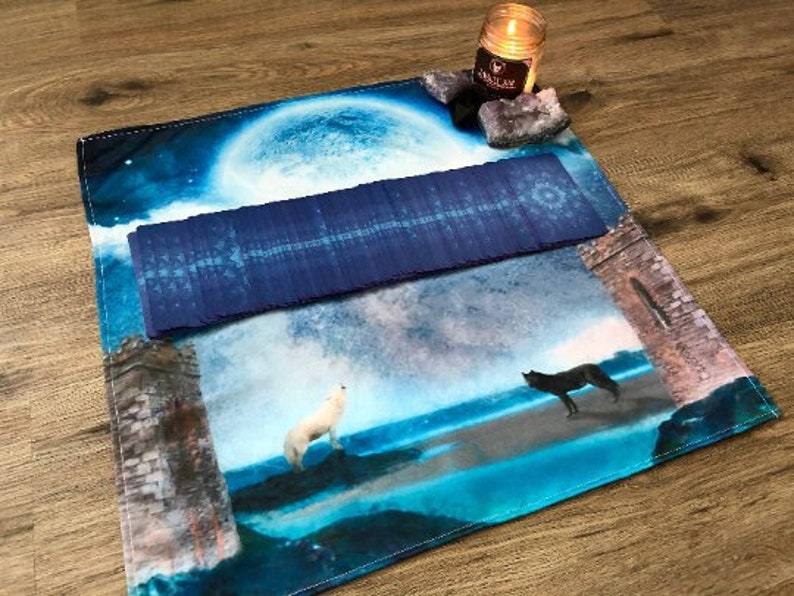 May include: A blue and white tarot cloth with a full moon, two wolves, and a stone archway. A deck of tarot cards is laid out on the cloth.