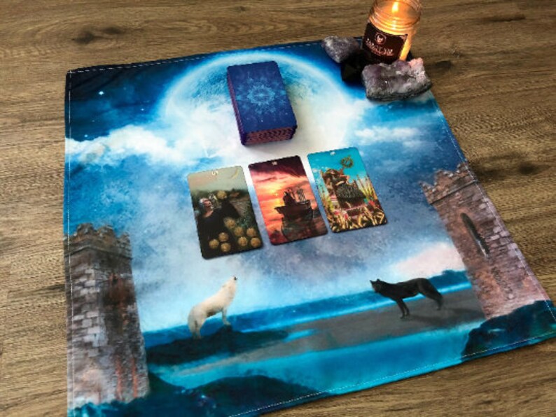 May include: A blue and white tarot cloth with a full moon, clouds, and two towers. Four tarot cards are laid out on the cloth, and a white wolf and a black wolf are howling at the moon. There is a candle and crystals on the top right corner of the cloth.