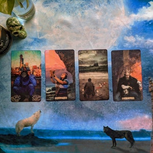 May include: Four tarot cards laid out on a blue and white patterned surface. The cards are "The Tower", "Queen of Cups", "Five of Swords", and "King of Swords". The cards are surrounded by a white candle, a skull, and two wolves.
