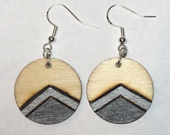 Könnte beinhalten: Ein Paar Holzohrringe mit einem silbernen geometrischen Design. Die Ohrringe sind rund und haben ein silbernes Chevron-Muster auf einem hellbraunen Holzgrund.
