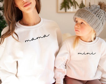 May include: A white sweatshirt with the word "mama" written in cursive on the front. A child is wearing a white sweatshirt with the word "mini" written in cursive on the front.