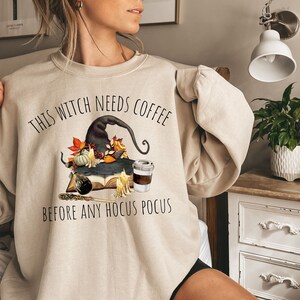 Könnte beinhalten: Ein beiger Pullover mit einem Hexenhut, einer Kaffeetasse und dem Text "This Witch Needs Coffee Before Any Hocus Pocus".