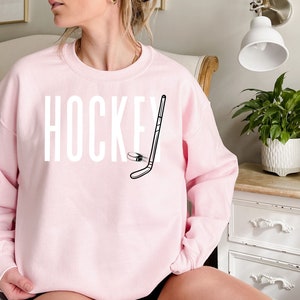 May include: A pink sweatshirt with the word "HOCKEY" printed in white and a black and white illustration of a hockey stick and puck.