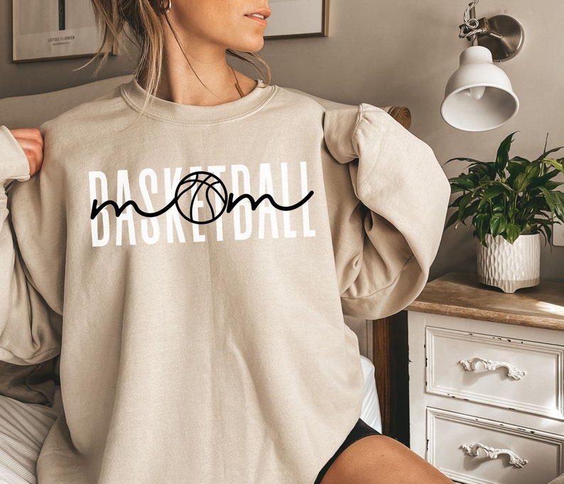 Puede incluir: Una sudadera beige con el texto "Basketball Mom" en letras blancas y un gr&aacute;fico de baloncesto negro.