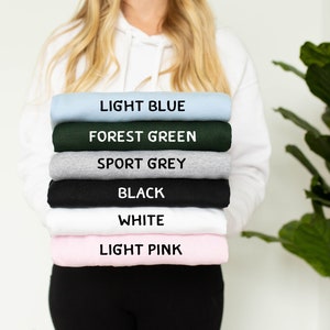 May include: A stack of six sweatshirts in different colors: light blue, forest green, sport grey, black, white, and light pink. The sweatshirts are folded and stacked on top of each other.