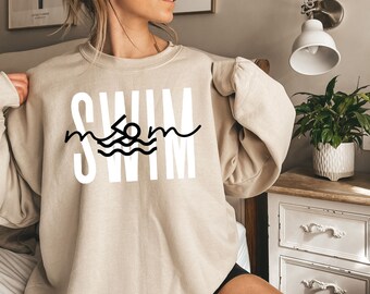 Könnte beinhalten: Ein beiger Pullover mit dem Wort "SWIM" in weißen Buchstaben bedruckt. Das Wort "SWIM" ist mit einem schwarzen Umriss und einer kleinen schwarzen Illustration einer Person, die in Wellen schwimmt, gestaltet.
