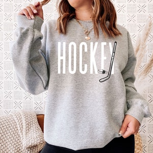May include: Gray sweatshirt with the word "HOCKEY" printed in white and a black and white illustration of a hockey stick and puck.