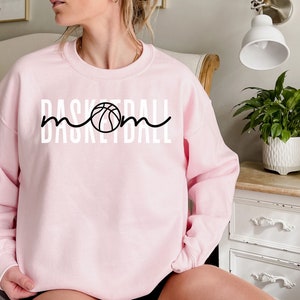 Puede incluir: Sudadera rosa con un gr&aacute;fico blanco que dice "Basketball Mom" con un gr&aacute;fico de baloncesto en el medio de la palabra "Mom".