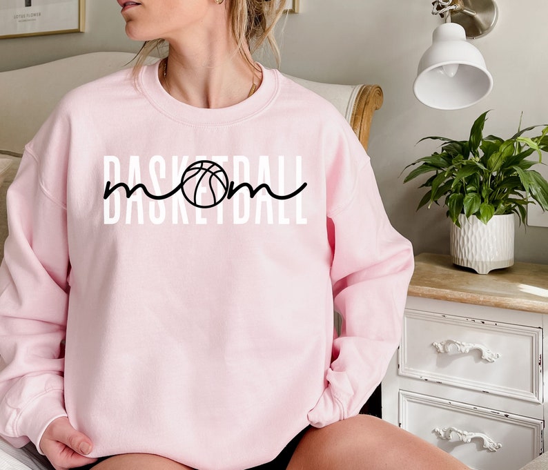 Puede incluir: Sudadera rosa con un gr&aacute;fico blanco que dice "Basketball Mom" con un gr&aacute;fico de baloncesto en el medio de la palabra "Mom".