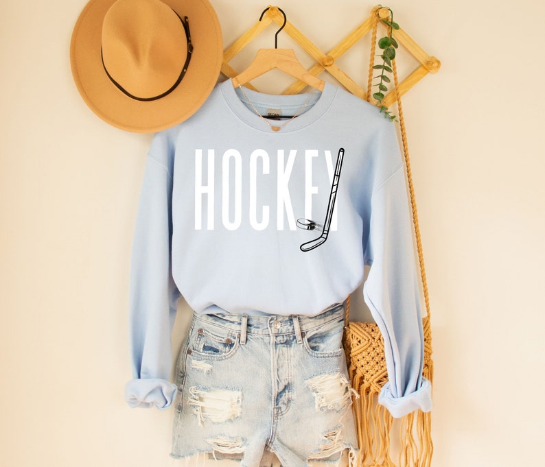 May include: Light blue sweatshirt with the word "HOCKEY" in white text and a black and white hockey stick and puck graphic.
