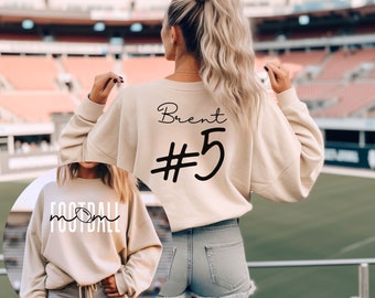 May include: A beige sweatshirt with the text "Brent #5" printed on the back. A second beige sweatshirt with the text "Football Mom" printed on the front. The sweatshirts are shown on a person wearing denim shorts.