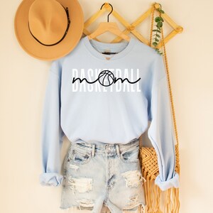 Puede incluir: Sudadera azul claro con el texto "Basketball Mom" en blanco y un gr&aacute;fico de baloncesto negro.