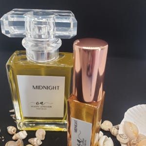 May include: Two bottles of perfume oil, one in a clear glass bottle with a gold cap and the other in a smaller bottle with a rose gold cap. Both bottles have a white label with the text "MIDNIGHT" and "OA OASIS ATELIER PERFUME OIL". The bottles are sitting on a bed of seashells.