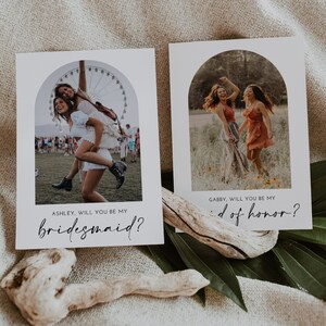 May include: Two cards with photos of two women each. The text on the cards reads "Ashley, will you be my bridesmaid?" and "Gabby, will you be my maid of honor?"