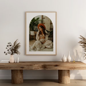 May include: A framed photograph of a pregnant woman wearing a white dress and a red hat, sitting in a room with plants. The photo is in a wooden frame with a white mat and an arched opening.