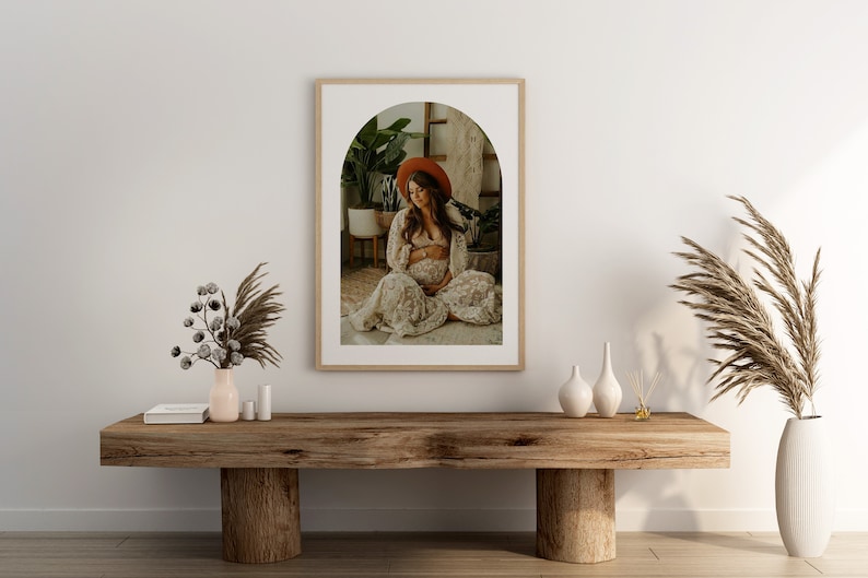 May include: A framed photograph of a pregnant woman wearing a white dress and a red hat, sitting in a room with plants. The photo is in a wooden frame with a white mat and an arched opening.