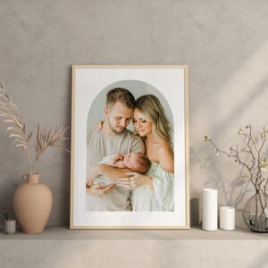 May include: A framed photograph of a family of three. A man and woman are holding a newborn baby in their arms. The photo is in a light wood frame with a white mat. The background is a soft, neutral color.