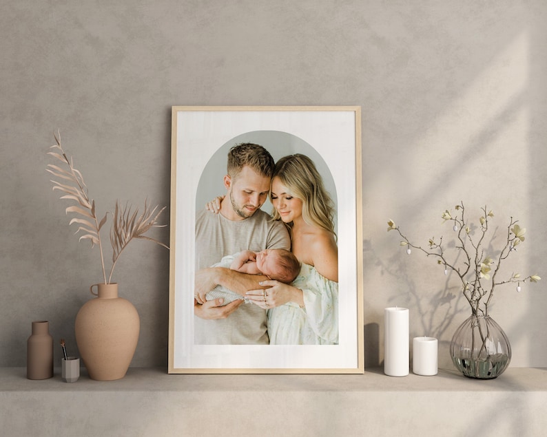 May include: A framed photograph of a family of three. A man and woman are holding a newborn baby in their arms. The photo is in a light wood frame with a white mat. The background is a soft, neutral color.