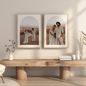 May include: Two framed photographs of a couple in a desert landscape. The photos are in a minimalist style with a white mat and a natural wood frame. The couple is wearing white clothing and is standing in a desert landscape with a large archway in the background.