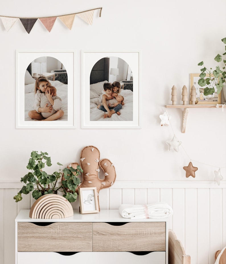 May include: Two framed photos of children in a nursery setting. The photos are in white frames with an arched opening. The photos are hung on a white wall with a white wainscoting paneling. The photos show children hugging and smiling.
