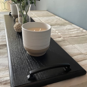 May include: A long, rectangular black wooden tray with two black metal handles on the ends. The tray holds a white ceramic candle with horizontal ridges, a clear glass vase containing artificial flowers, and two small decorative white spheres.