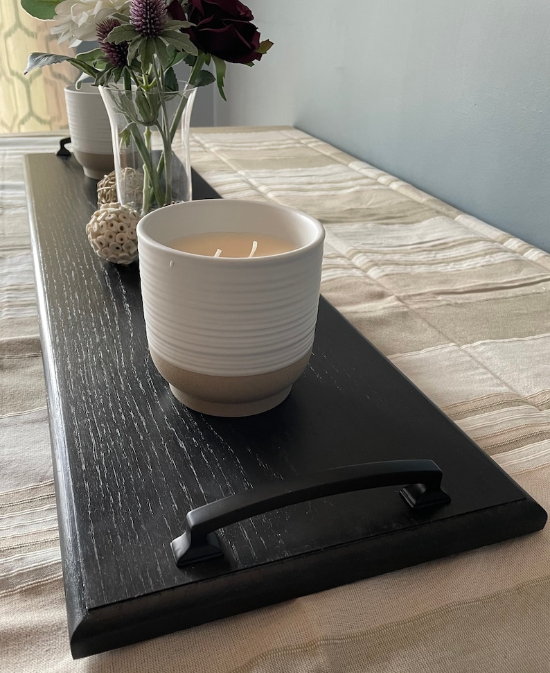 May include: A long, rectangular black wooden tray with two black metal handles on the ends. The tray holds a white ceramic candle with horizontal ridges, a clear glass vase containing artificial flowers, and two small decorative white spheres.