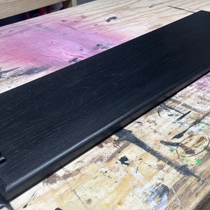 May include: A long, rectangular wooden serving tray finished in a deep black stain that highlights the natural wood grain. The tray features two curved, matte black metal handles securely attached to each end of the flat, elongated surface.