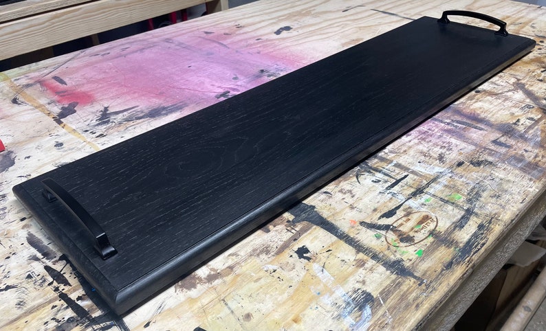 May include: A long, rectangular wooden serving tray finished in a deep black stain that highlights the natural wood grain. The tray features two curved, matte black metal handles securely attached to each end of the flat, elongated surface.