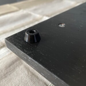 May include: A black wooden board featuring a smooth, dark finish with a single conical black rubber foot attached to the corner. The surface shows a visible wood grain texture, and a small metal screw head is embedded nearby on the flat panel.