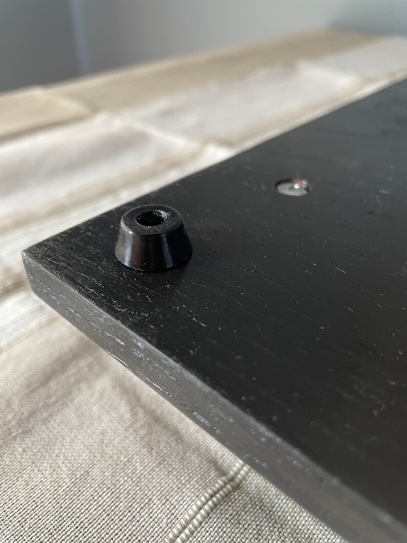 May include: A black wooden board featuring a smooth, dark finish with a single conical black rubber foot attached to the corner. The surface shows a visible wood grain texture, and a small metal screw head is embedded nearby on the flat panel.