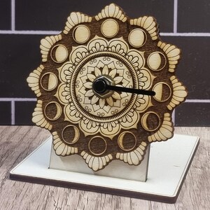 May include: A wooden clock with a mandala design and moon phases around the edge. The clock has a black hand and is mounted on a white stand. The clock is made of light brown wood and is a decorative item.