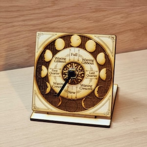 May include: A wooden desk clock featuring a detailed depiction of the phases of the moon. The clock face is circular, with the phases labeled and a sunburst design at the center. The clock is square and stands on a wooden base.