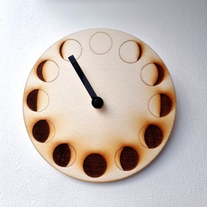 May include: A wooden clock with a black hand and a moon phase design. The clock face is divided into 12 sections, each representing a different phase of the moon.