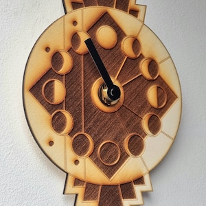 May include: A wooden clock with a circular face featuring a moon phase design. The clock has a black hour hand and a gold minute hand. The clock is mounted on a white wall.